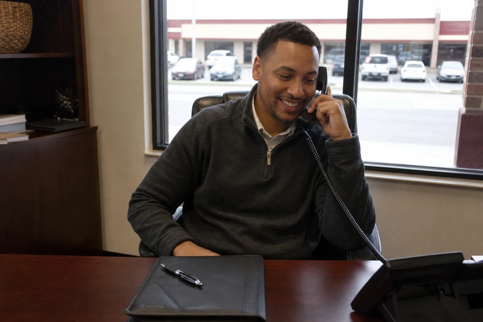 Smiling agent in a gray pullover talks on a desk phone at a wooden desk with a black folder and pen, a window to a strip-center parking lot behind him